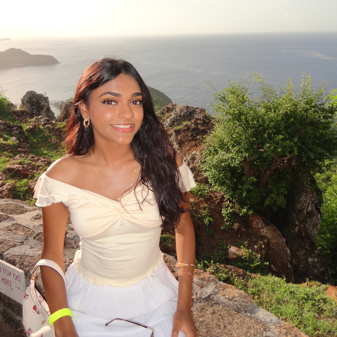 A woman smiling while sitting on a stone ledge with a scenic ocean and rocky landscape in the background.