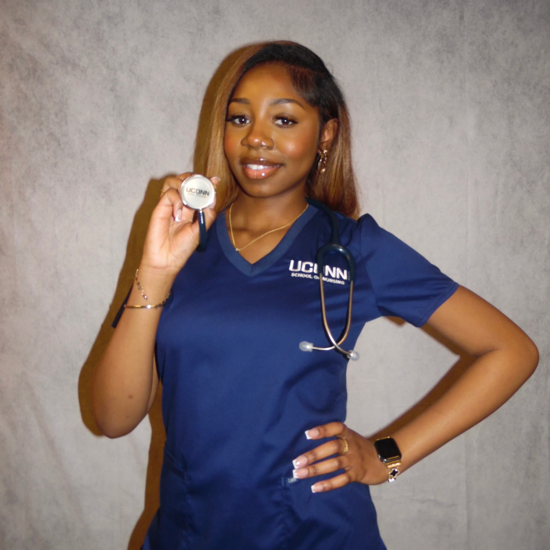 A woman in navy blue scrubs holding a stethoscope and smiling, standing against a plain background.