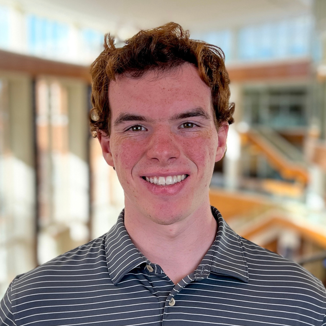 A young man smiling indoors, wearing a striped polo shirt, with a bright and modern building interior in the background.