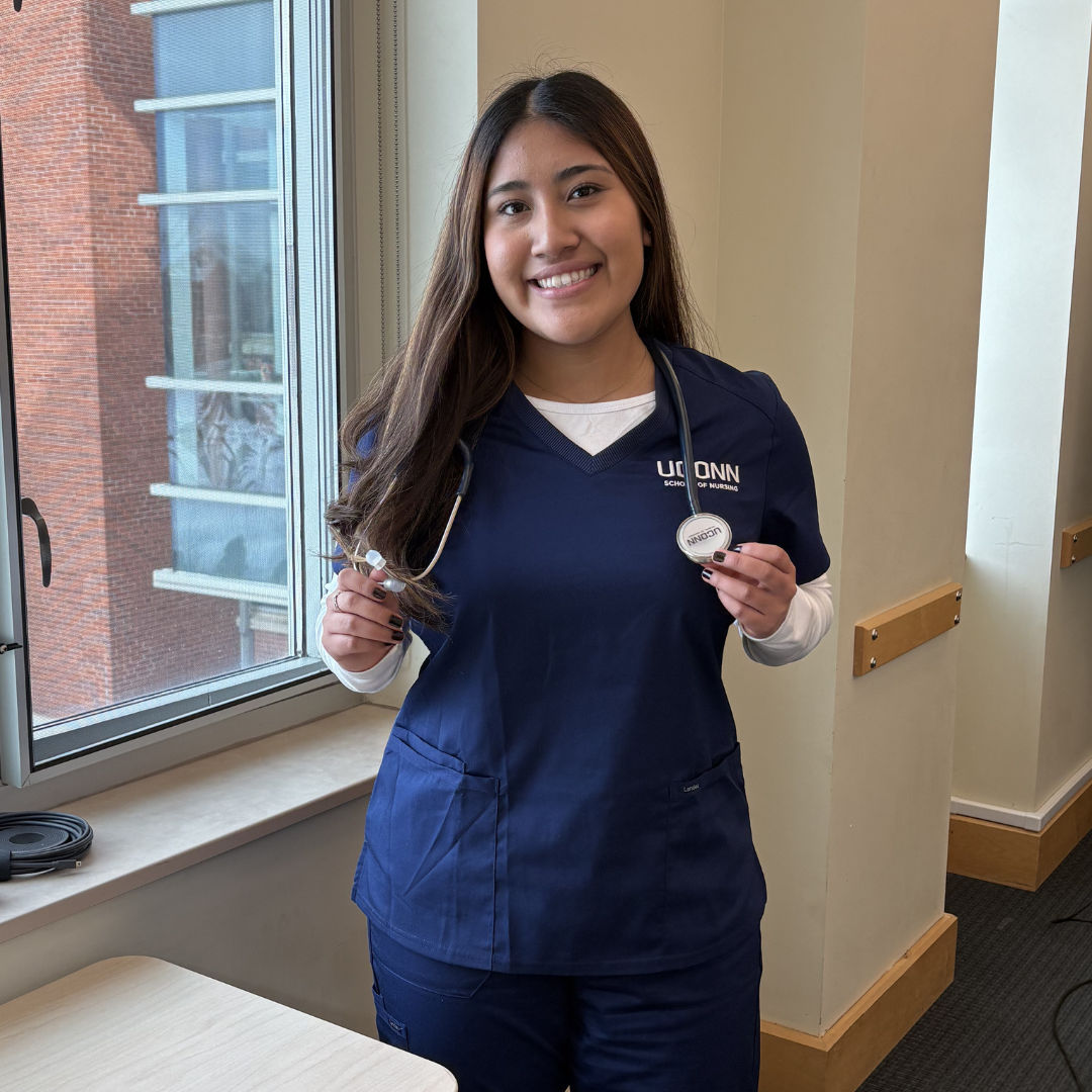 A woman in navy blue scrubs smiling and holding a stethoscope, standing by a window in a bright indoor setting.