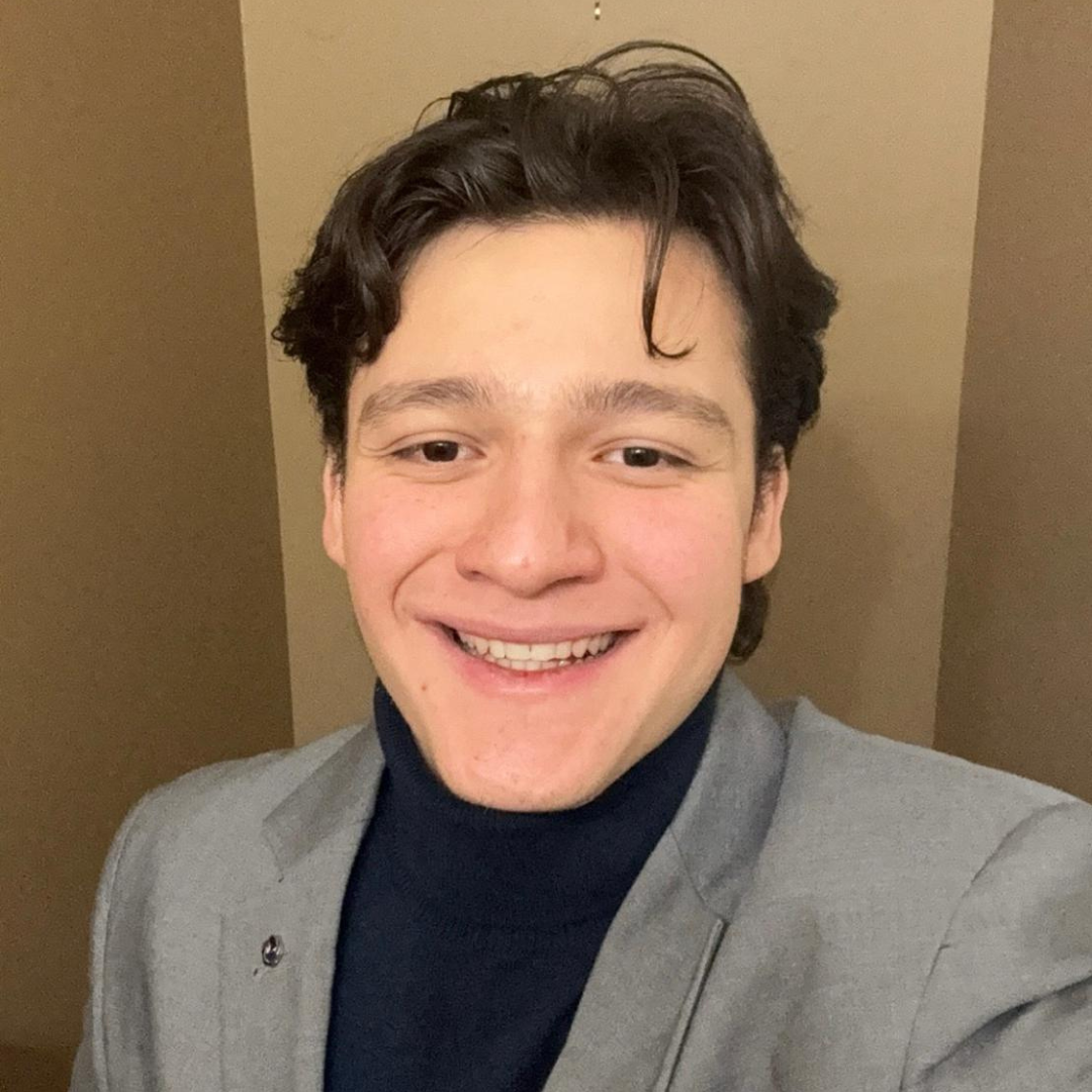 Smiling man with dark hair wearing a gray blazer and dark turtleneck, posed against a beige background.