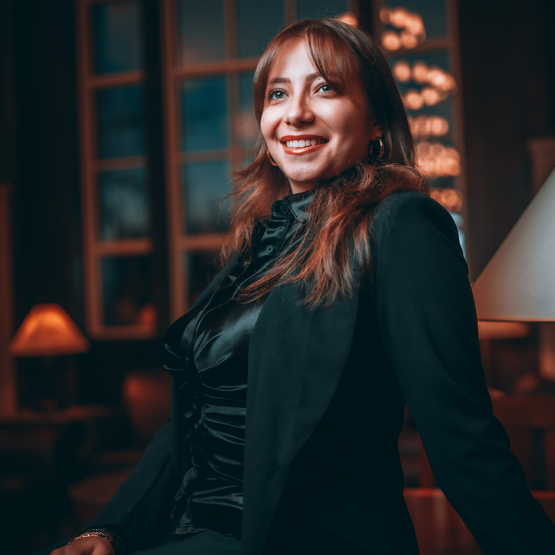 A woman smiling and posing indoors in a dimly lit, elegant setting, wearing a black outfit and blazer.