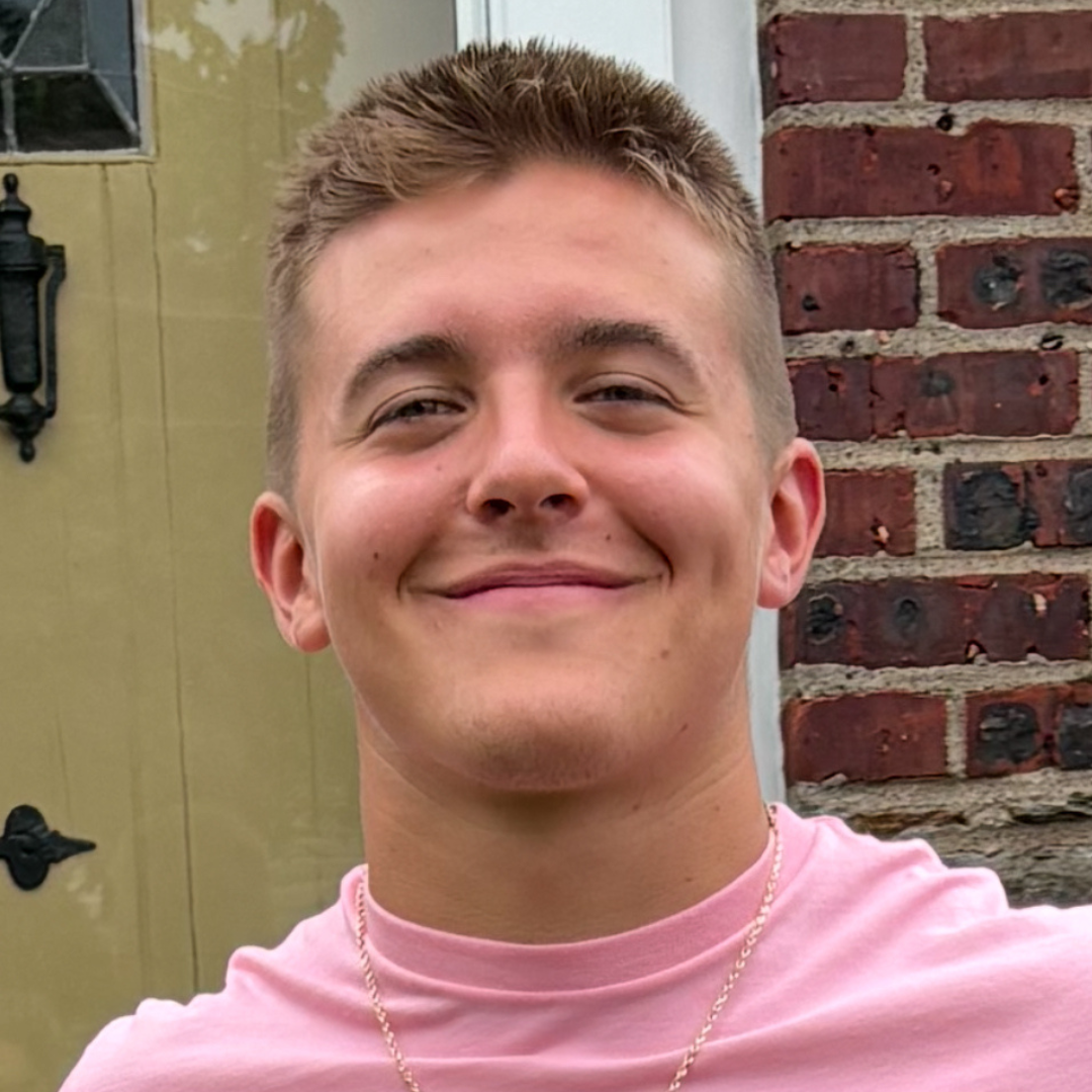 A young man smiling outdoors, wearing a pink shirt and standing in front of a brick wall and door.