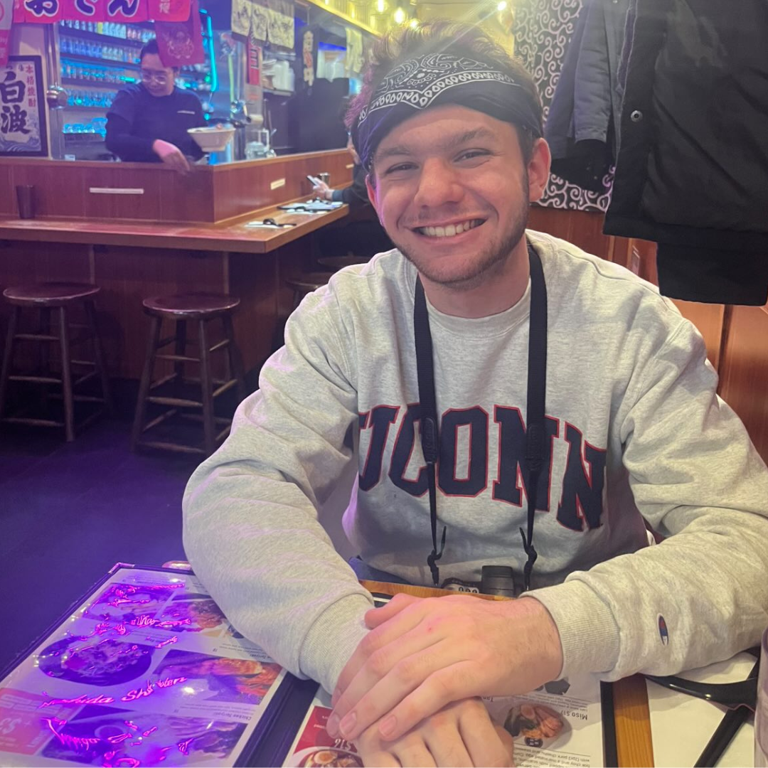 A man smiling at a table in a lively restaurant, wearing a UConn sweatshirt and a bandana.
