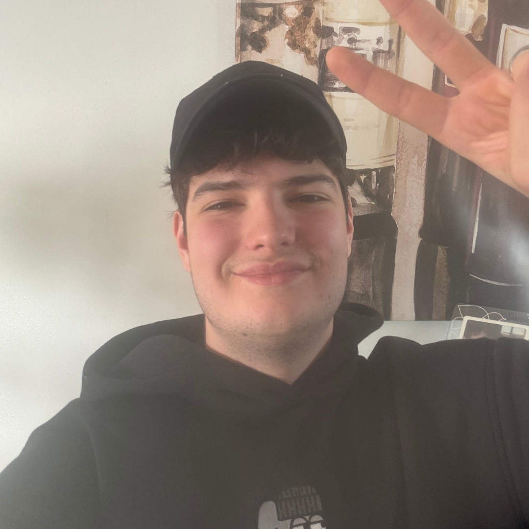 A young man smiling and holding up a peace sign, wearing a black hoodie and cap indoors.