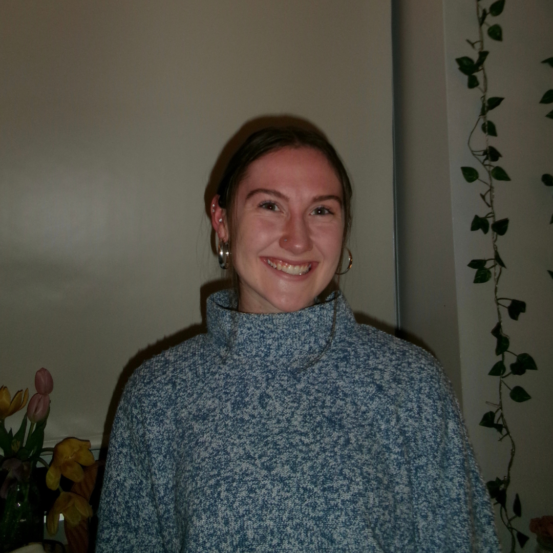 A woman smiling indoors, wearing a blue sweater with plants and flowers in the background.