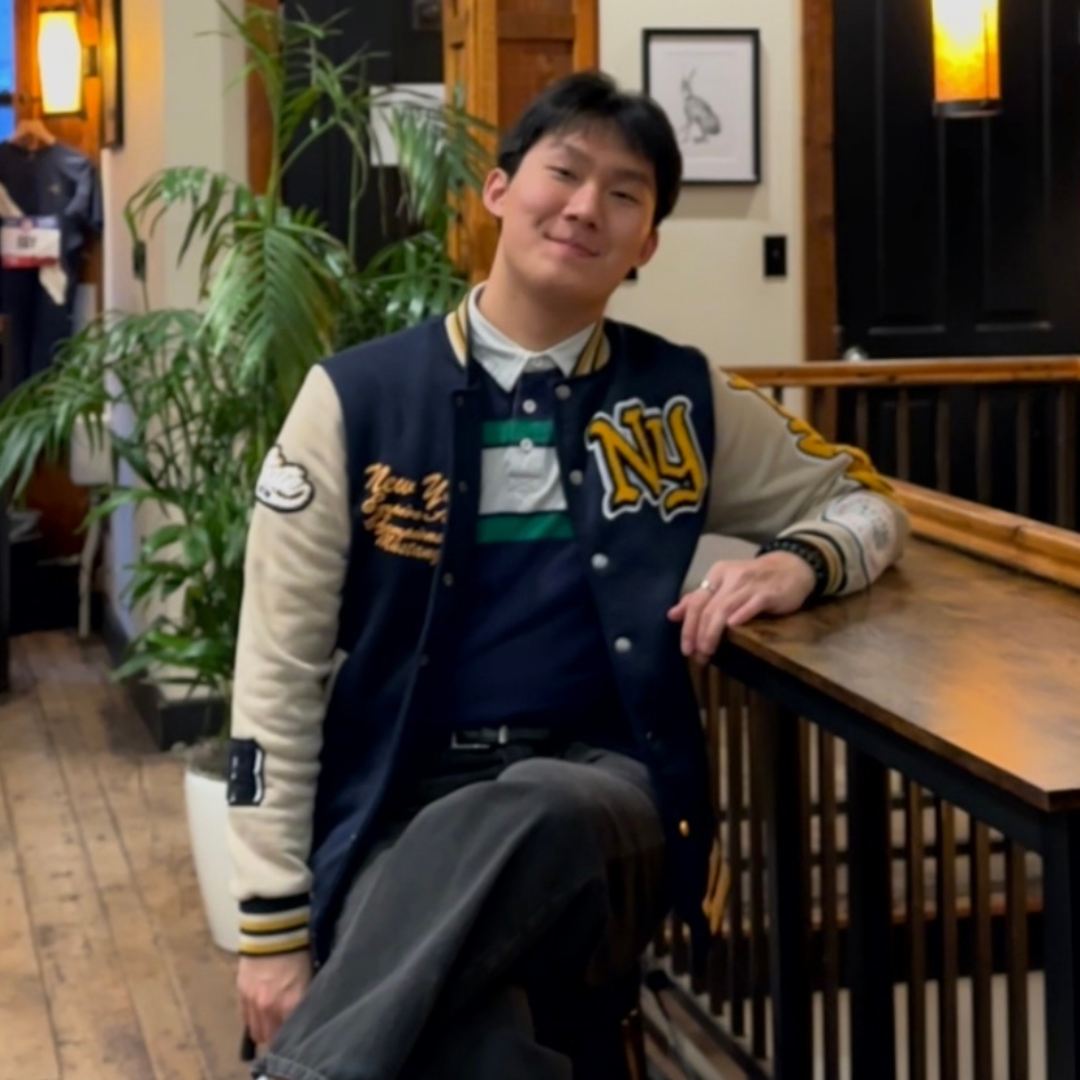 A man smiling and leaning on a wooden railing indoors, wearing a varsity jacket and sitting in a warmly lit space with plants and artwork in the background.