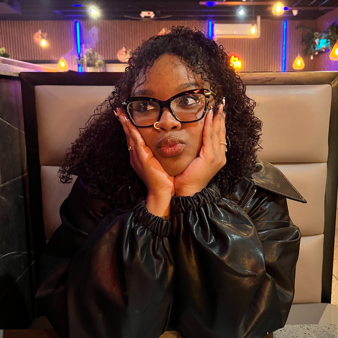  A woman with curly hair and glasses resting her face in her hands, seated in a booth at a warmly lit restaurant.