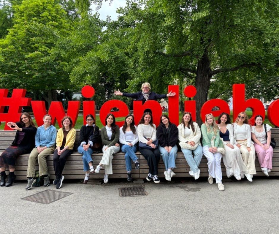 A group of thirteen students and one instructor from the EGL program sit and pose cheerfully in front of a large red "#wienliebe" sign on a wooden bench in a park in Vienna, Austria. The instructor stands behind the sign with arms outstretched. The group is surrounded by lush green trees, highlighting the outdoor setting and cultural immersion experience.