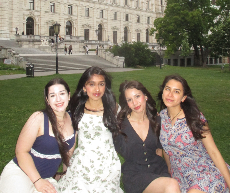 Four EGL students sit on a green lawn in front of the historic Hofburg Palace in Vienna, Austria. They are dressed in summer outfits and smiling at the camera, enjoying the cultural experience and peaceful outdoor setting. The ornate white facade of the palace, with arched windows and classical architectural details, rises in the background, adding a sense of place and history.
