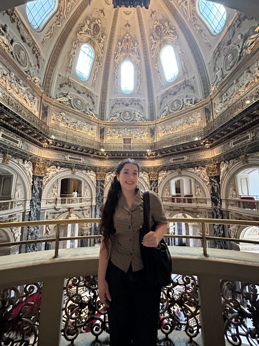 A young woman with long dark hair stands smiling inside a grand, ornately decorated building with high arched windows and an elaborately sculpted dome ceiling. She wears a checkered blouse, dark pants, and carries a black shoulder bag. Behind her, intricate architectural details, marble columns, and gold accents fill the museum-like space.
