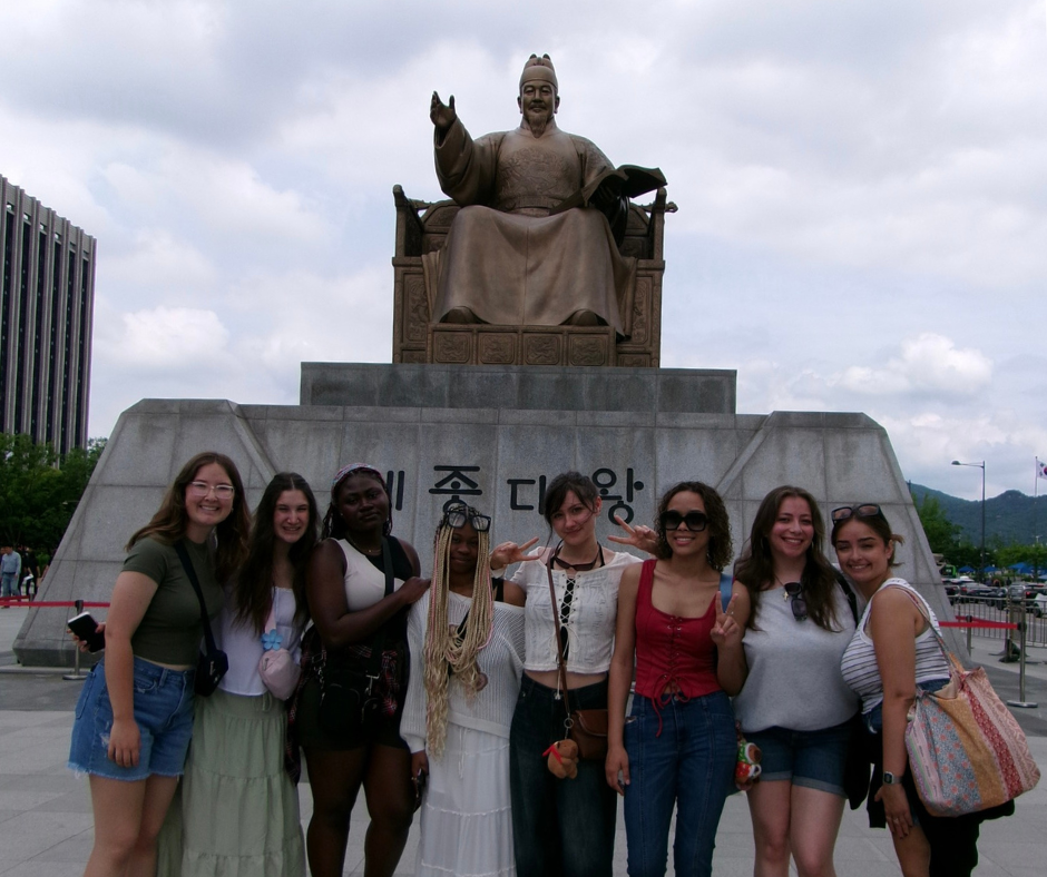 A diverse group of UConn students poses and smiles in front of the large bronze statue of King Sejong the Great in Seoul, South Korea. The statue sits atop a stone pedestal engraved with Korean text. The students appear cheerful, holding peace signs and wearing casual summer outfits during their cultural excursion.