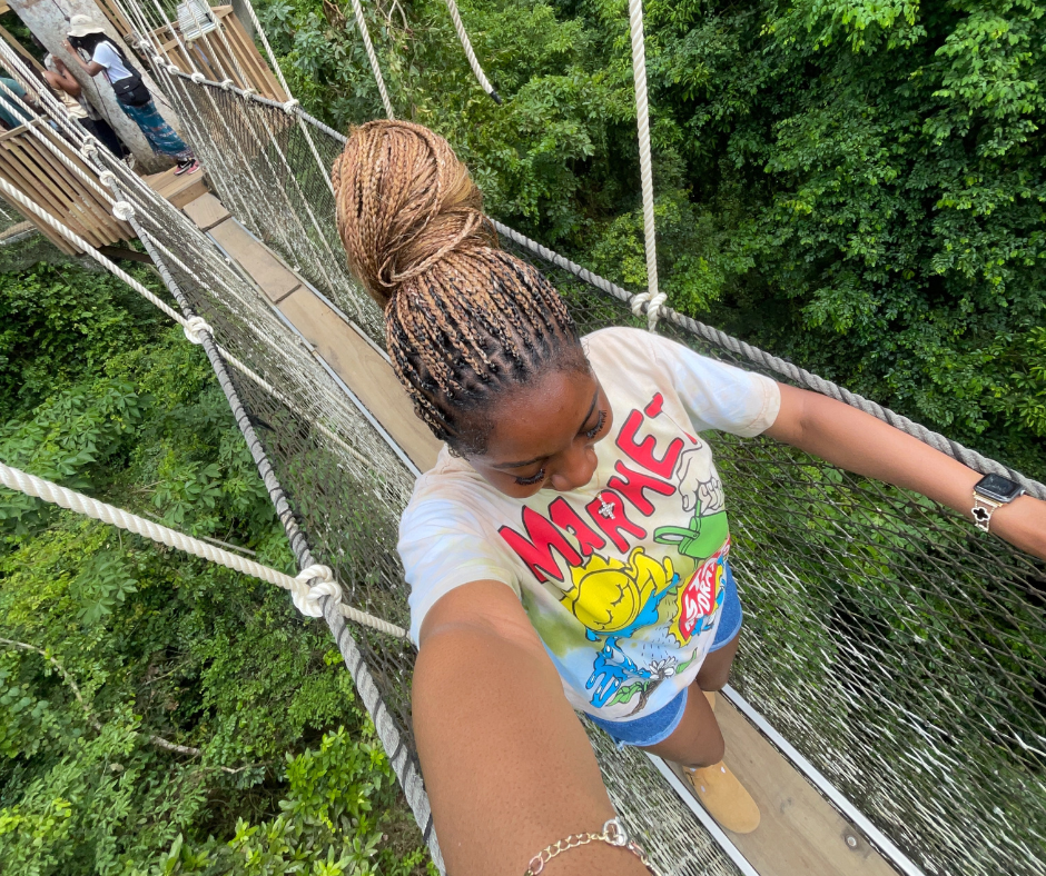 A young woman takes a selfie while walking on a narrow rope bridge suspended above a lush green forest. She wears a colorful graphic t-shirt, denim shorts, and boots, with her braided hair tied in a bun. The canopy walk is part of an adventurous experience in Ghana, highlighting outdoor exploration during her global journey.