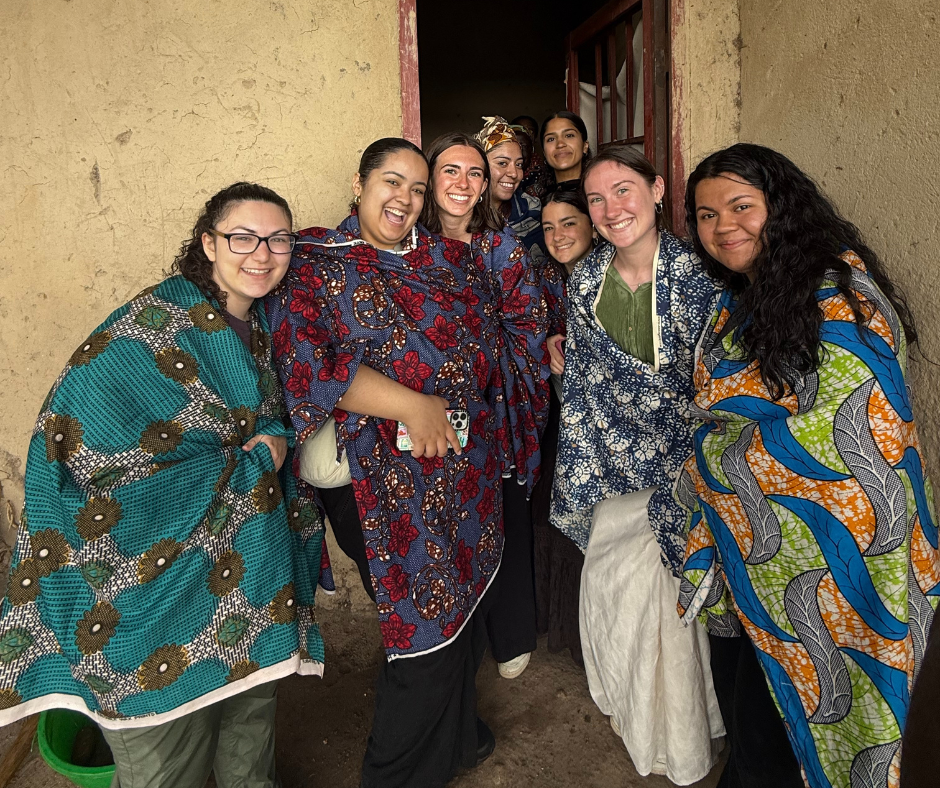 A group of smiling women stands together in front of a doorway, each wrapped in colorful traditional Rwandan fabric. The women are dressed in vibrant prints of blue, green, orange, and red, and appear joyful and connected. They pose in a rustic setting with a beige, textured wall behind them, suggesting an immersive cultural experience.