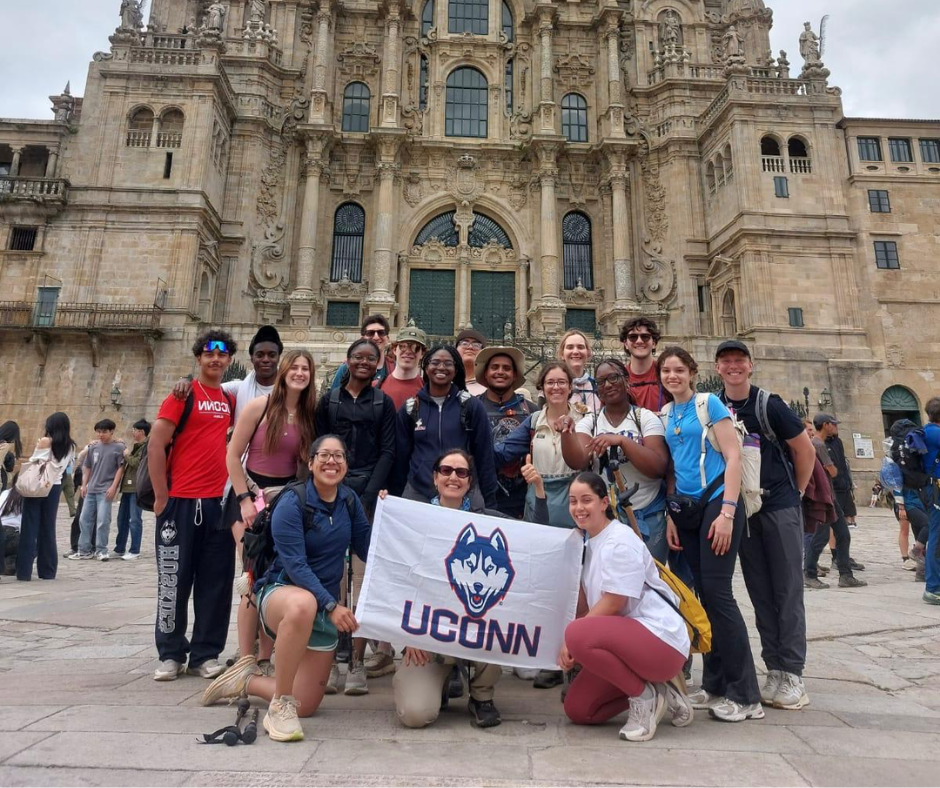 A diverse group of 18 UConn students smiling and posing together in front of an ornate historical building with stone architecture. Two students kneel in front holding a white UConn flag featuring a husky logo. The group wears casual travel attire and backpacks, suggesting they are on a study abroad trip.