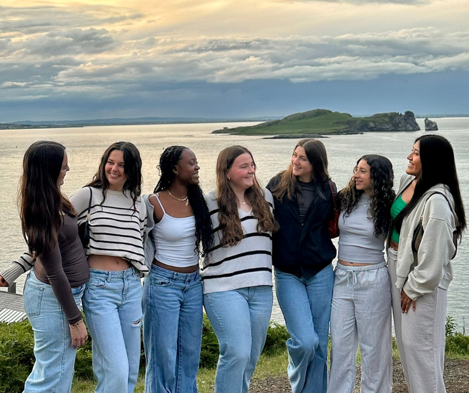 A group of seven young women standing outdoors in front of a scenic coastal view during sunset. They are smiling at each other with an island and calm water in the background.