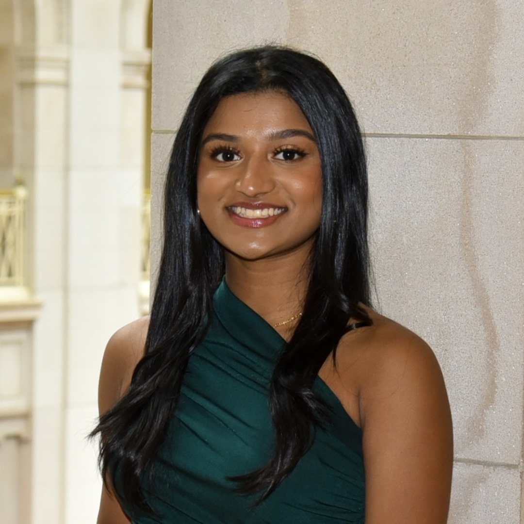 A young woman with long, straight black hair smiles at the camera while standing indoors in front of a stone wall. She is wearing a dark green, one-shoulder dress and a gold necklace. The background features a softly lit architectural setting with tall columns and gold railings, suggesting a formal or elegant environment.