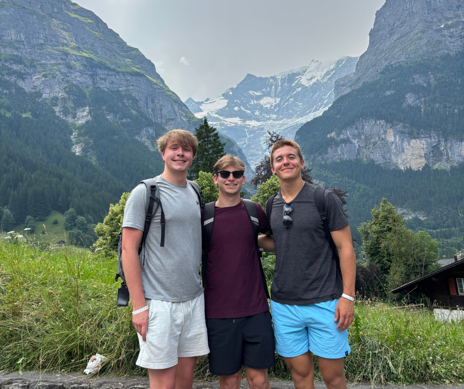  Three people stand arm-in-arm smiling in front of a scenic mountain landscape with green forests and snow-capped peaks in the background. All are wearing casual summer clothing and backpacks, suggesting they are hiking or sightseeing.