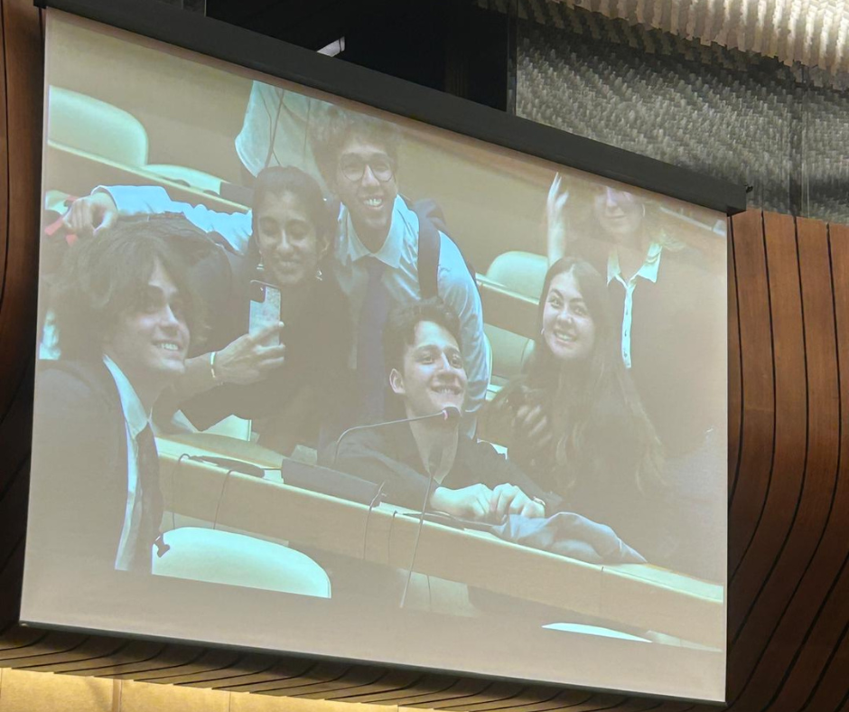 A projected image shows a group of six young adults dressed in business attire, smiling and taking a selfie together in what appears to be a formal conference room. They are seated at desks with microphones, and the atmosphere is cheerful and collegial.