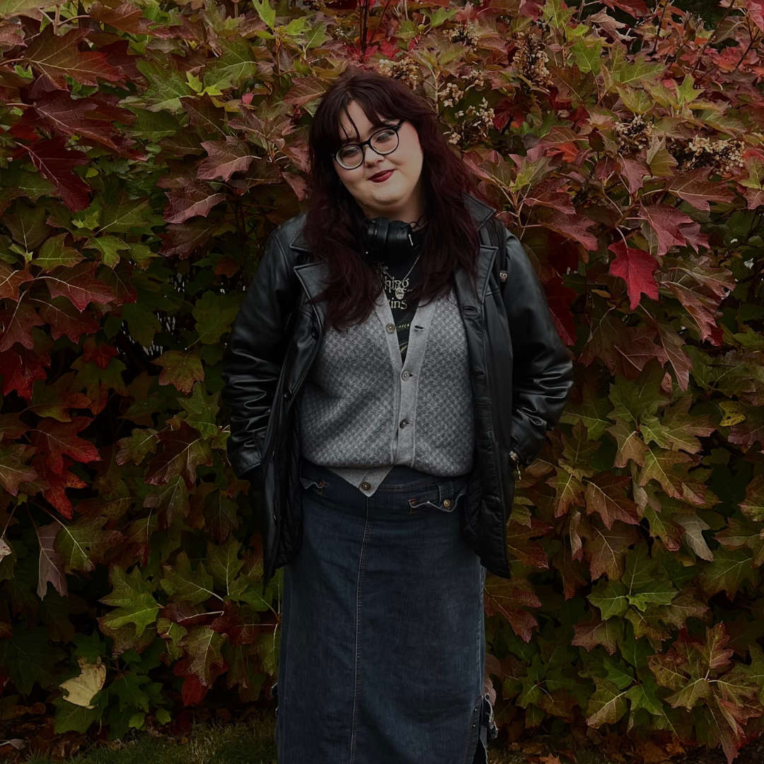A student stands in front of a backdrop of vibrant autumn leaves on the UConn campus. She is wearing a black leather jacket, gray buttoned cardigan over a graphic T-shirt, and a long denim skirt. She has dark red hair, glasses, and over-ear headphones around her neck. She is smiling with her hands in her jacket pockets, embodying a relaxed and confident fall campus vibe.