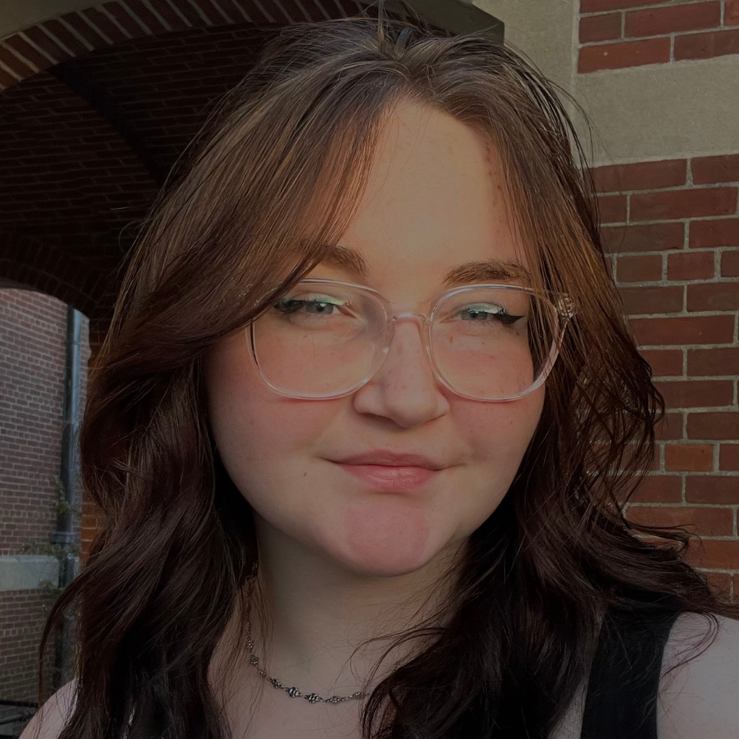 Close-up of a student standing in front of a red brick building on the UConn campus. She has wavy dark brown hair, clear-framed glasses, and is wearing a subtle chain necklace. Soft sunlight highlights her face as she smiles slightly, conveying a calm and confident presence.