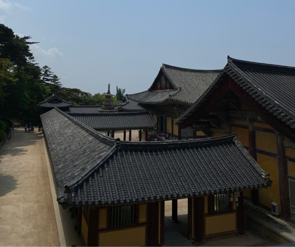 A scenic view of a traditional Korean temple complex featuring curved tiled rooftops, wooden beams, and earth-toned walls. The surrounding area includes a stone path and green trees under a clear blue sky. This photo captures a moment of cultural exploration at a historic site during a UConn Global experience abroad.