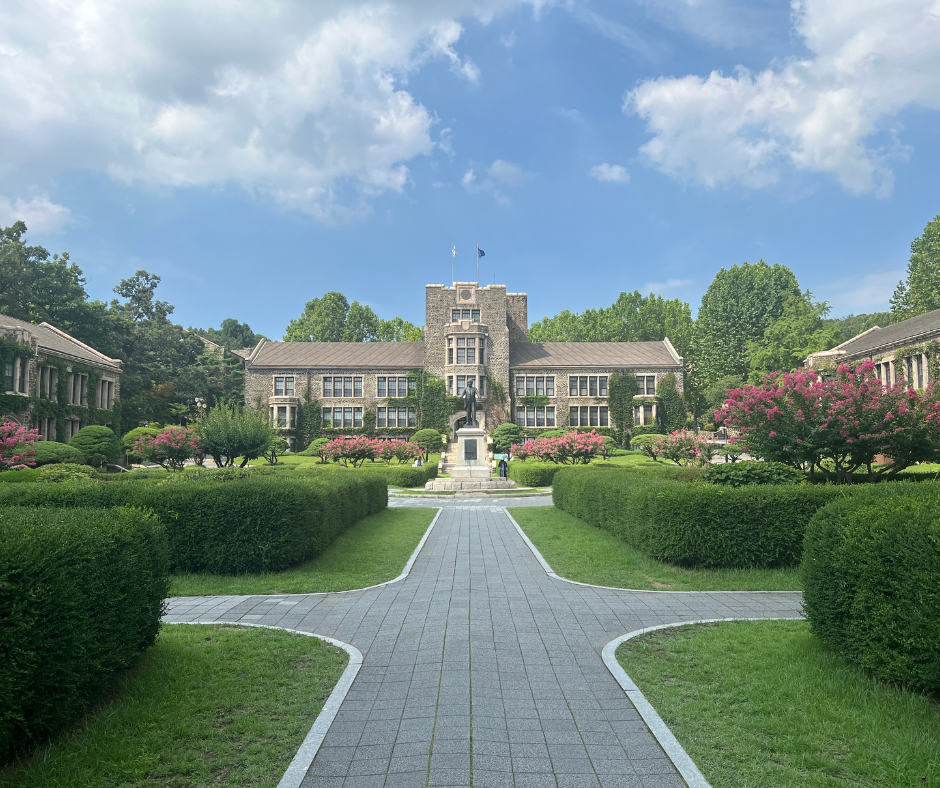 A panoramic view of a university campus in South Korea, featuring a grand stone building framed by symmetrical gardens and manicured hedges. A statue stands in the center of the walkway leading to the entrance. Pink flowering trees and a vibrant blue sky with scattered clouds create a welcoming and peaceful academic environment, reflecting the beauty of international study destinations available through UConn Global.