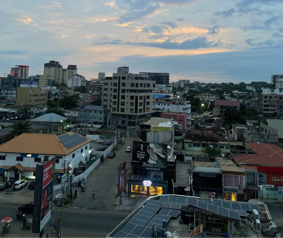 A wide view of Accra, Ghana at dusk, showing a mix of mid-rise buildings, shops, and residences. The sky is painted with soft hues of orange and blue as the sun sets, and city lights begin to flicker on. Rooftop solar panels and street-level signs are visible, showcasing the urban energy of the capital city.