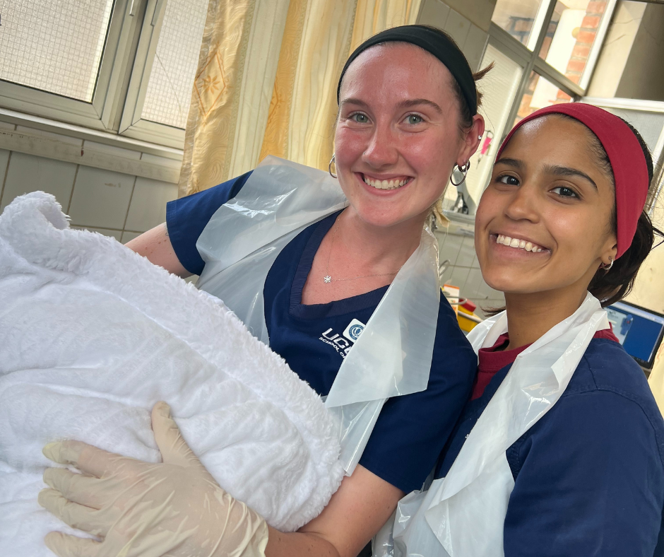 Two young women in navy blue scrubs and plastic aprons smile in a brightly lit hospital room. One is holding a newborn baby wrapped in a white blanket, while the other stands close beside her. Both are wearing headbands and gloves, showcasing their involvement in hands-on nursing work during a clinical experience in Rwanda