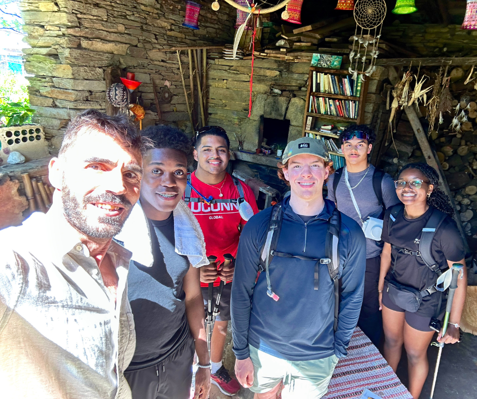 A group of six UConn students and one guide inside a rustic stone-walled room decorated with colorful textiles, dreamcatchers, and bookshelves. The students wear hiking gear and backpacks, holding walking sticks, and are smiling for the photo, indicating they are engaged in an outdoor adventure or cultural immersion activity abroad.