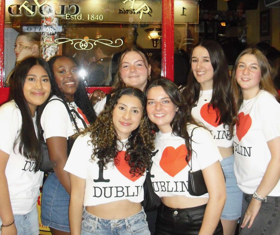 Seven young women posing together at night in front of a pub window. They are all wearing matching "I ♥ Dublin" t-shirts and smiling at the camera.