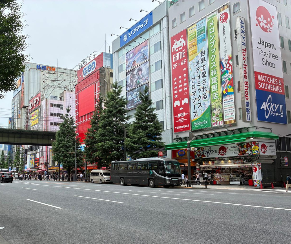 A busy street in Akihabara, Tokyo, Japan, lined with colorful buildings covered in large advertisements and signage. A black tour bus with "Grand Limousine Tokyo" written on the side is parked near a tax-free souvenir shop. Pedestrians can be seen in the distance, and traffic flows beneath a green railway bridge overhead. Tall green trees line both sides of the wide street, adding contrast to the urban scenery.