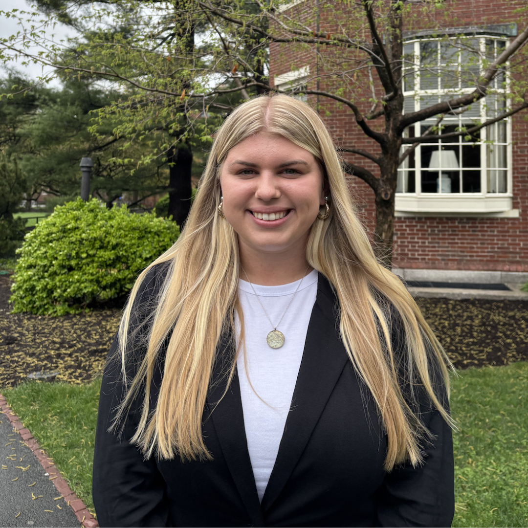 A young woman with long, straight blonde hair stands outdoors, smiling at the camera. She is wearing a black blazer over a white top and a round pendant necklace. Behind her is a red brick building with a large white-trimmed window, surrounded by green trees, a lawn, and a path lined with bricks. The scene suggests a campus or formal garden setting on a cloudy day.