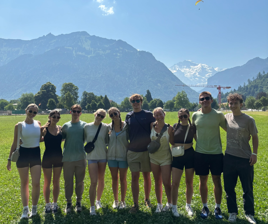 A group of ten people stand together in a grassy field with their arms around each other, smiling at the camera. Behind them are tall mountains, including snow-covered peaks, with a paraglider visible in the sky. The group is dressed in casual summer attire.