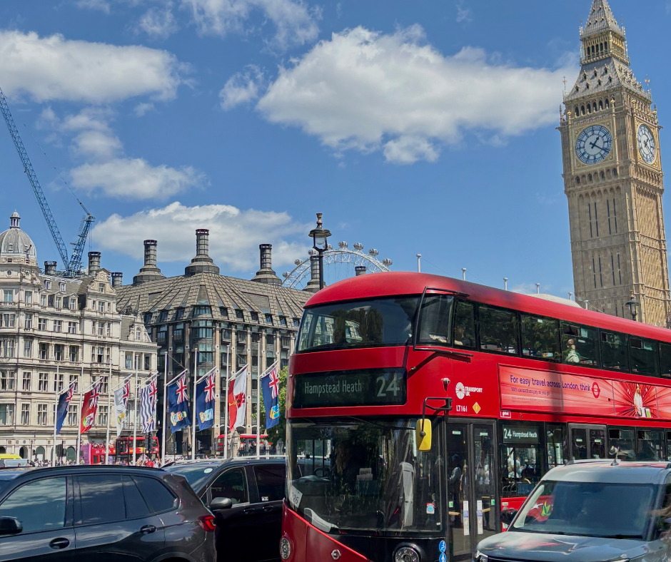 A red double-decker bus drives through central London with the Big Ben clock tower and UK flags in the background. A bright blue sky with scattered clouds highlights the iconic cityscape.