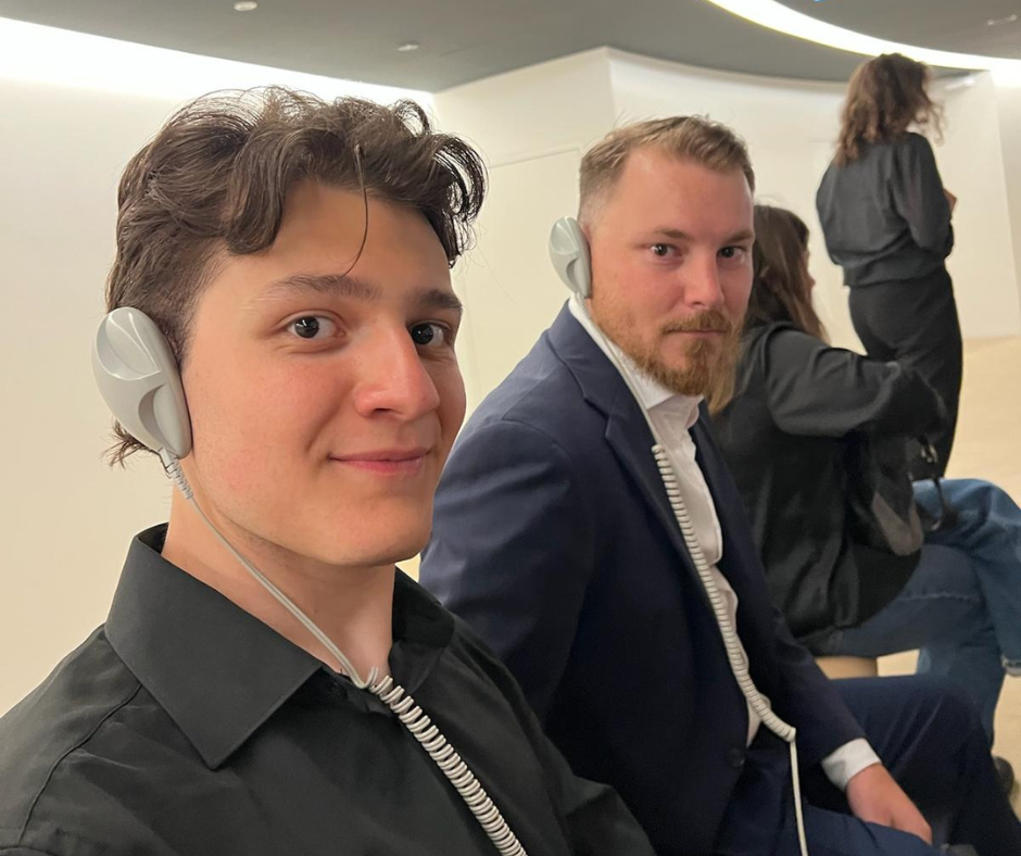 Two men sit side by side wearing translation headsets in a modern conference space. The man on the left has short dark hair and a black shirt, smiling slightly at the camera. The man on the right has light hair, a beard, and is dressed in a navy suit jacket with a white shirt, looking directly at the camera. Other attendees are visible in the background.