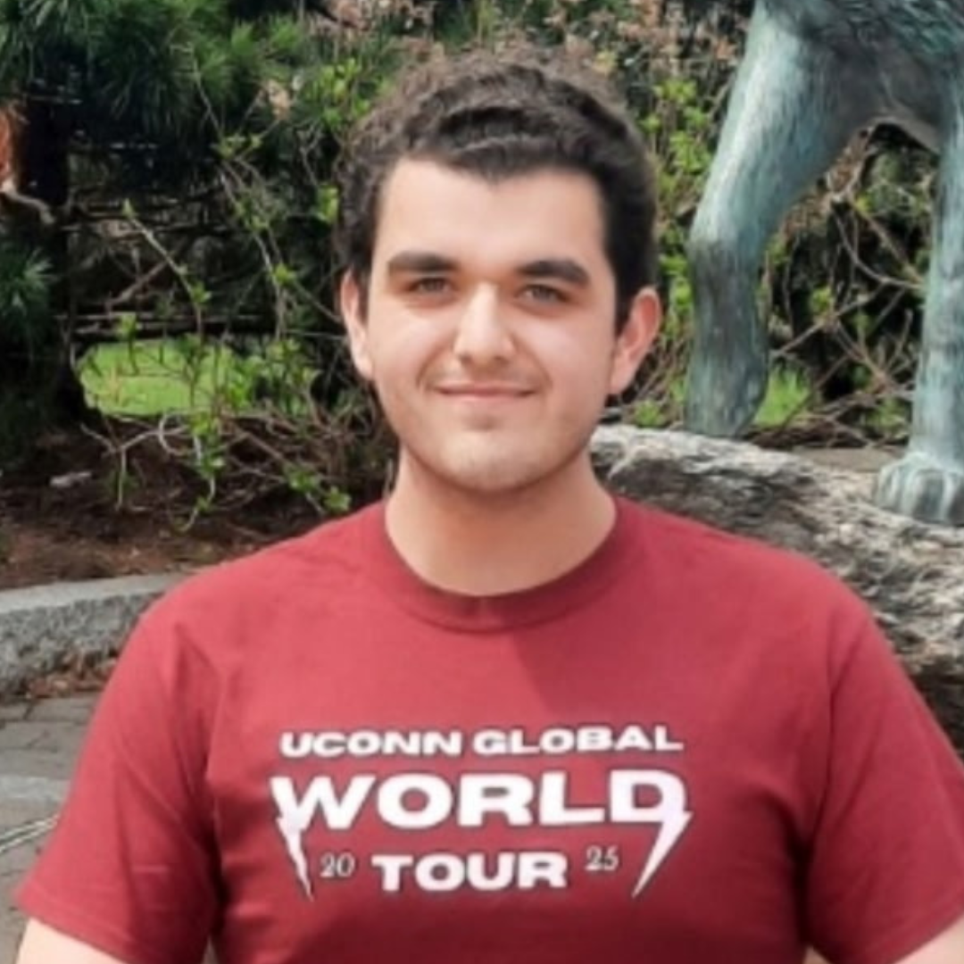 A young man with short, dark curly hair smiles at the camera while standing outdoors. He is wearing a maroon t-shirt that reads "UConn Global World Tour 2025" in bold white letters with a lightning bolt graphic. In the background, there is greenery and part of a large statue, likely depicting an animal's leg, along with stone landscaping, suggesting a campus or park setting.