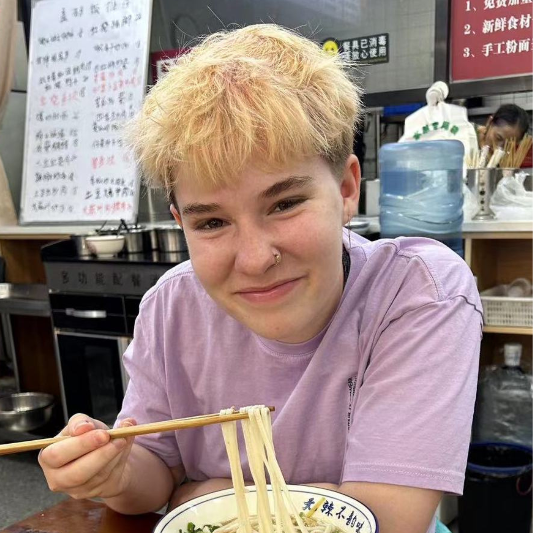  A young person with short, blond hair and a nose ring smiles while holding chopsticks with noodles in a casual noodle shop. They are wearing a lavender T-shirt and seated at a wooden table with a bowl of noodles in front of them. Behind them are handwritten menus in Chinese and a large water jug. The setting appears to be a local eatery, suggesting a cultural or international dining experience.