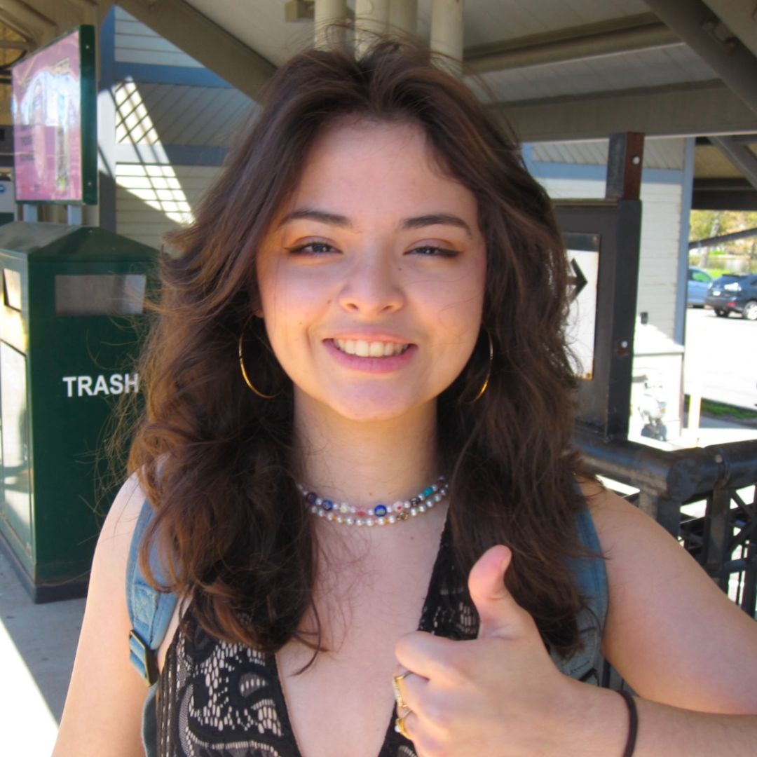 Alt text: A young woman with long, wavy brown hair smiles and gives a thumbs-up while standing outdoors. She is wearing hoop earrings, layered beaded necklaces, and a sleeveless patterned top with a backpack over her shoulders. The background includes a green trash bin labeled "TRASH" and part of a building structure, indicating a public or campus setting on a sunny day.