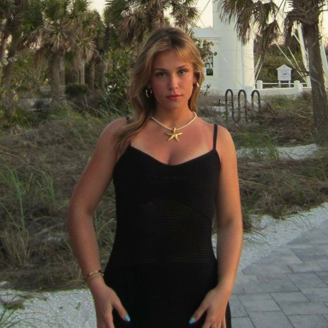 Alt text: A young woman stands confidently on a paved path near a beach, with palm trees and a white building in the background. She is wearing a black sleeveless dress, a gold starfish necklace, hoop earrings, and has long, wavy blonde hair. The natural lighting suggests it is either early morning or late afternoon. The setting evokes a calm, coastal atmosphere.