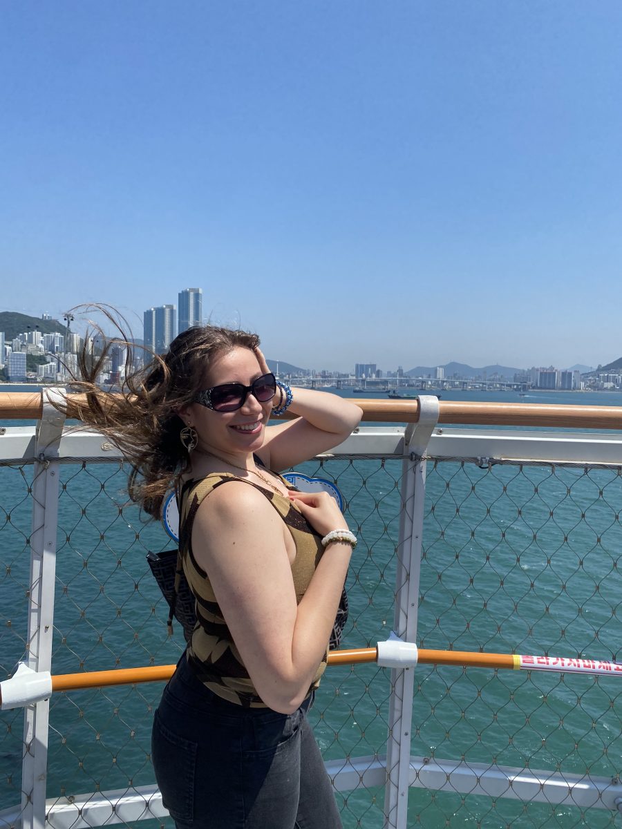 A young woman smiles while standing on a boat deck with the ocean and a city skyline in the background. Her hair blows in the wind as she poses in front of a safety railing with netting. She wears sunglasses, a sleeveless top, dark jeans, and a backpack, enjoying a sunny day during a global experience abroad.
