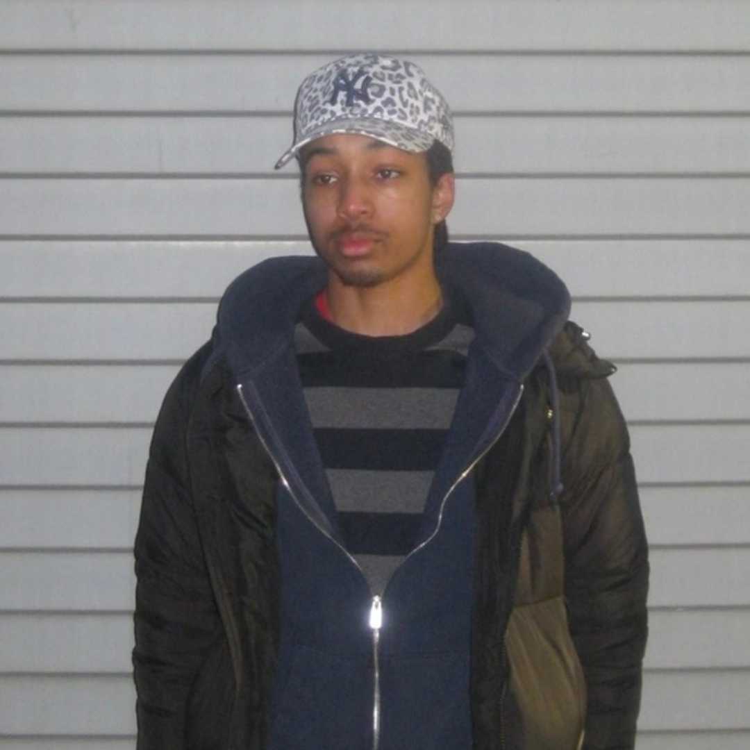 Alt text: A young man stands in front of a light gray, horizontally paneled wall. He is wearing a striped black and gray sweater, a navy blue zip-up hoodie, a black puffer jacket, and a white baseball cap with a gray leopard print pattern and a "NY" logo. His expression is neutral, and he is looking slightly away from the camera. The lighting suggests the photo was taken indoors or with a flash.