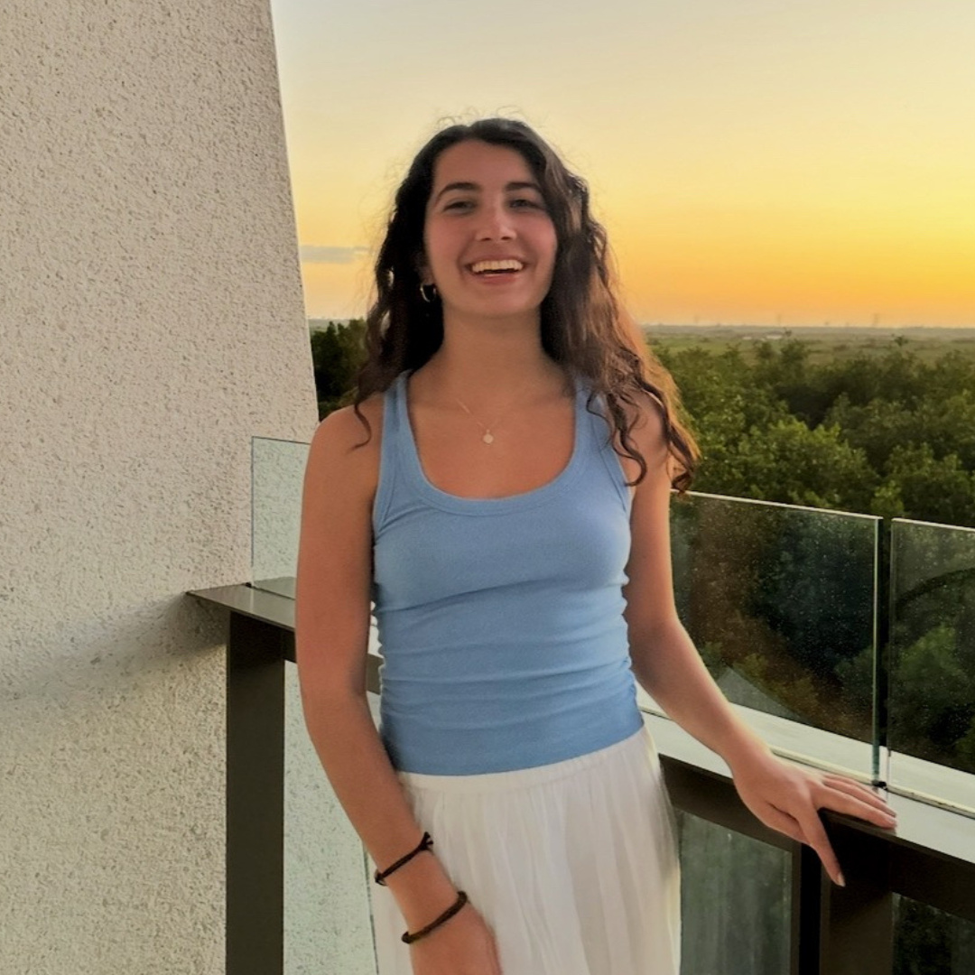 A young woman with long curly dark hair smiles while standing on a balcony at sunset. She wears a light blue sleeveless top and a white skirt. The balcony has a glass railing, and behind her, there is a view of green trees and a clear sky with warm orange and yellow tones from the setting sun.