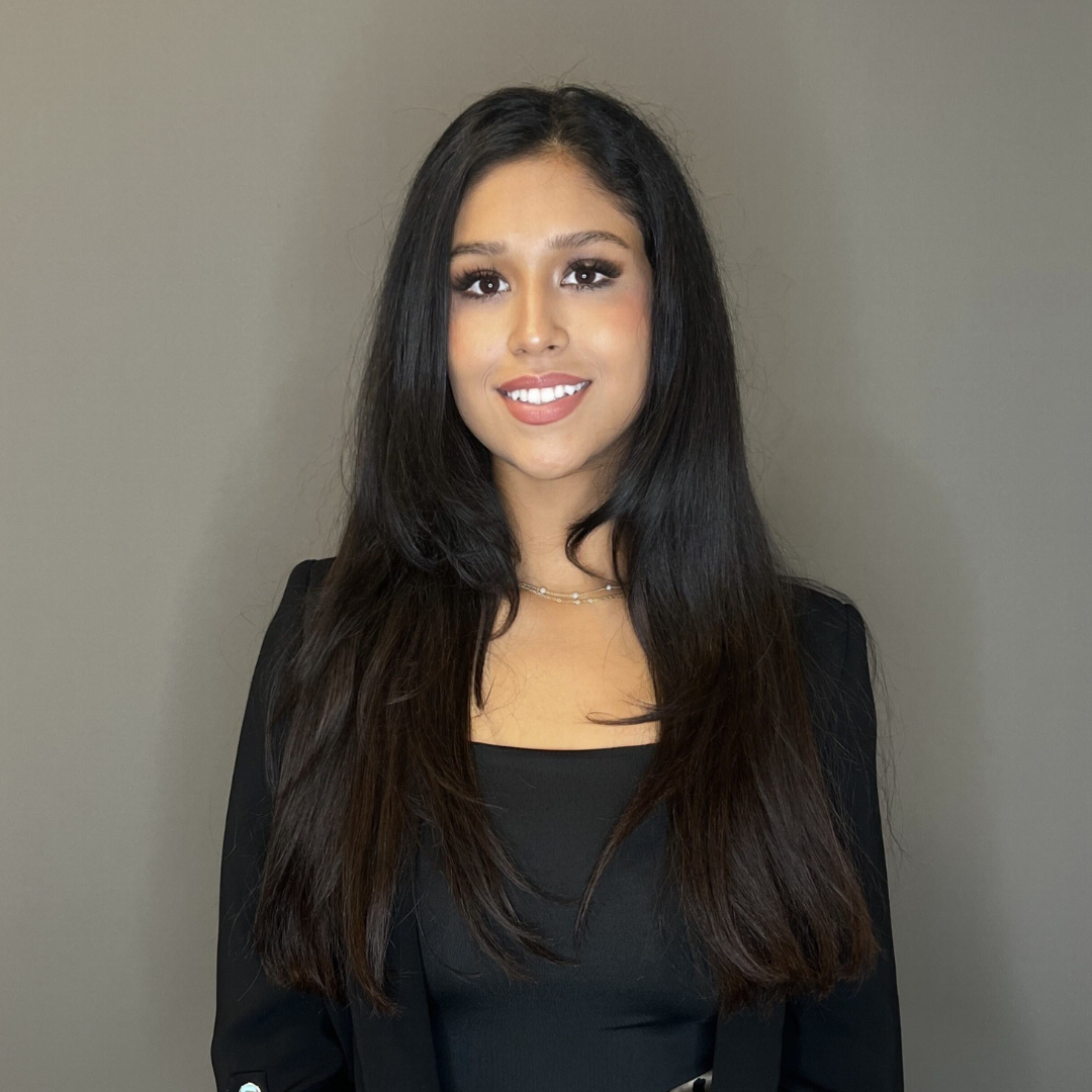 Portrait of a young woman with long, straight black hair wearing a black blazer and top, smiling against a plain gray background.