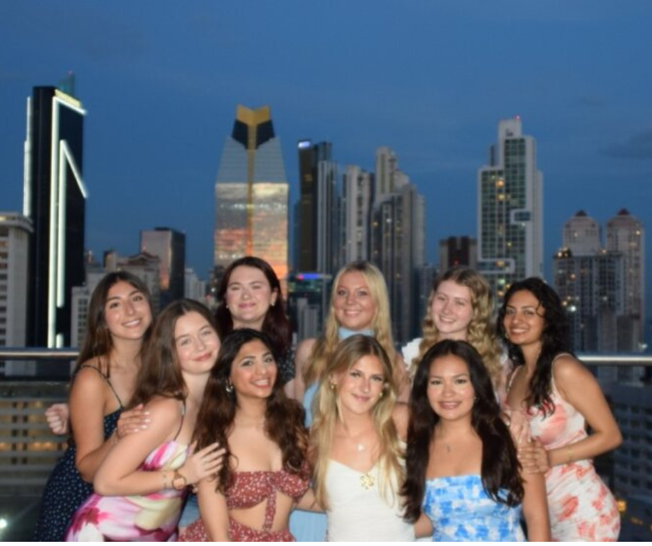 A group of ten young women pose together on a rooftop at dusk, with a modern city skyline featuring tall, illuminated skyscrapers in the background. They are dressed in colorful, summery dresses and are smiling warmly, creating a celebratory and joyful atmosphere against the urban evening backdrop.
