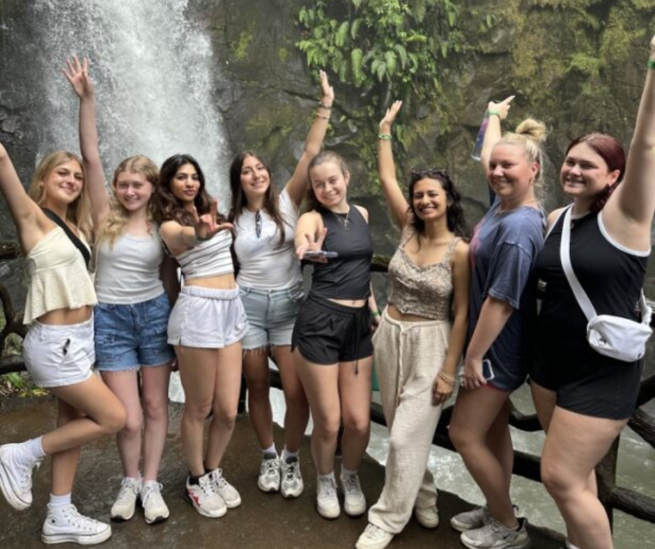 A group of eight young women pose cheerfully in front of a lush waterfall during an outdoor adventure. They are standing on a wet stone pathway with greenery and mist around them, wearing casual summer clothing and athletic shoes. Most have their arms raised or extended in joyful, celebratory gestures.