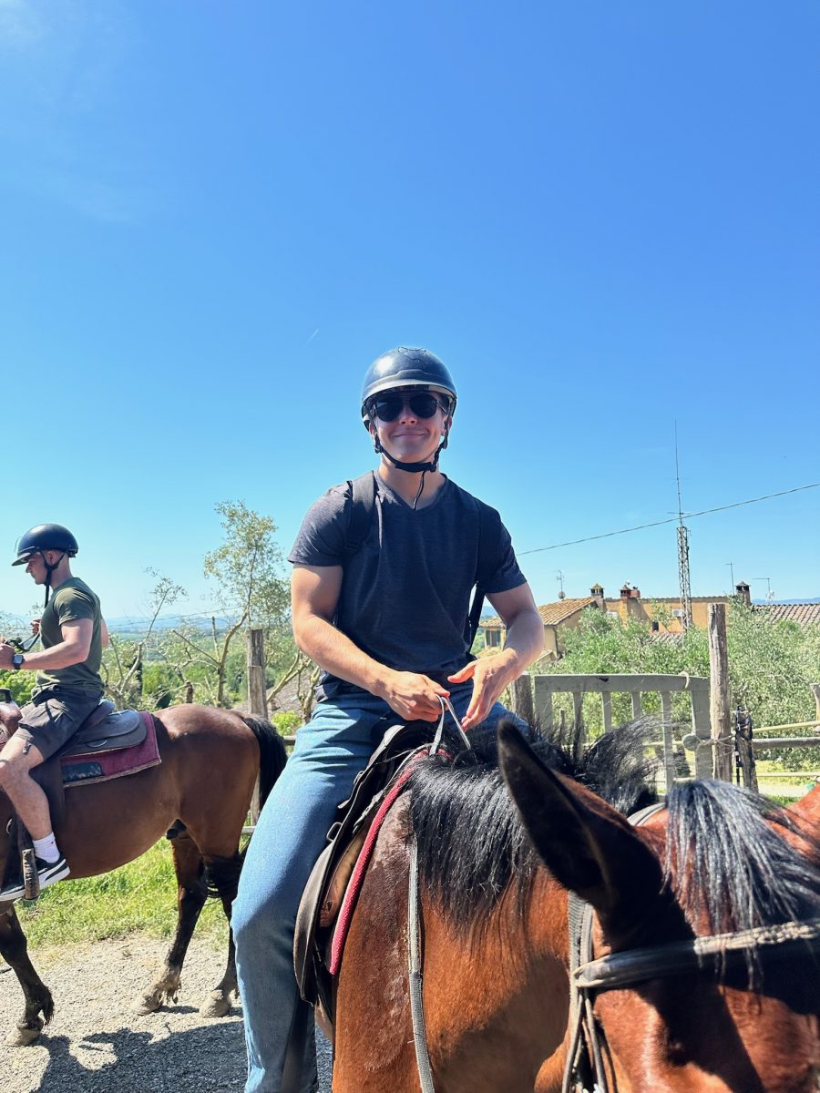 A person wearing a helmet, sunglasses, a dark t-shirt, and jeans is horseback riding on a sunny day. The horse has a brown coat with a dark mane. Another rider on a brown horse is visible in the background to the left. The scene includes a clear blue sky, wooden fencing, greenery, and rooftops in the distance.