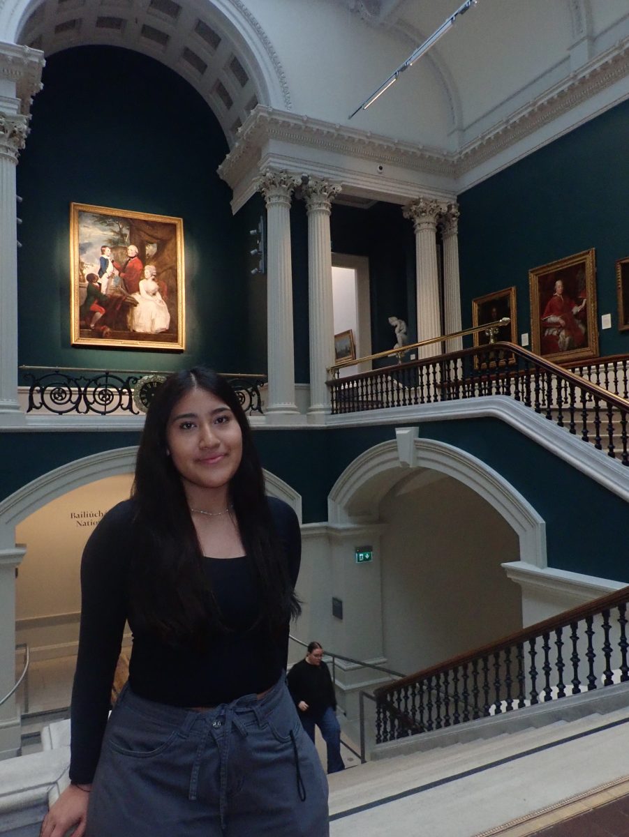 A young woman standing on a staircase inside an art museum with classical architecture, arched doorways, and large framed paintings on dark green walls.