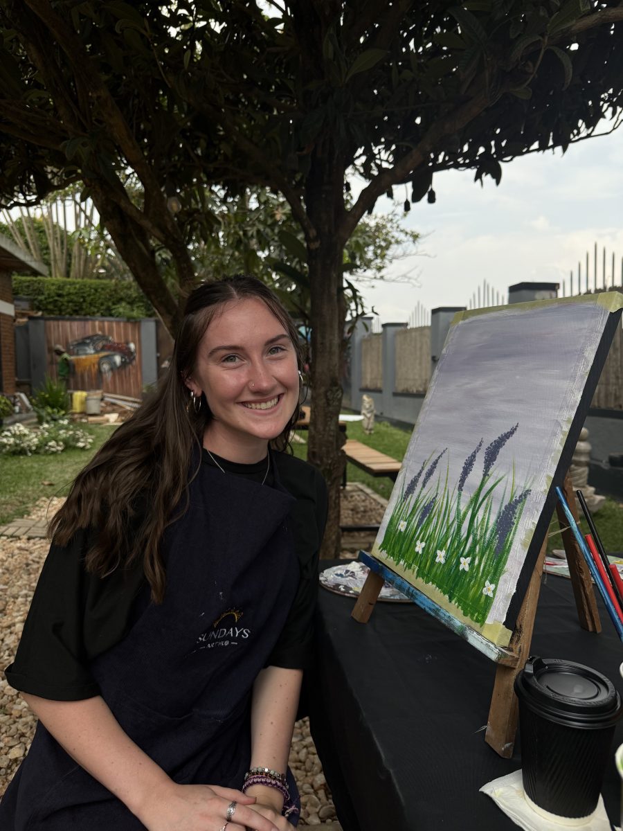 A young woman smiles while seated outdoors at a painting station, wearing a dark apron that reads “SUNDAYS KIGALI.” She sits beside an easel displaying her artwork of tall lavender flowers and white blossoms against a soft gray sky. The setting includes trees, a gravel path, and a peaceful garden area with greenery and painted walls in the background, capturing a creative moment during her study abroad experience in Rwanda.