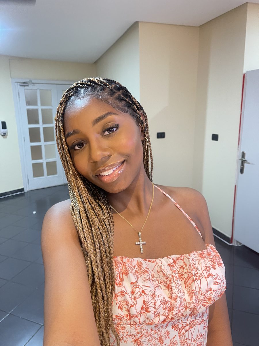 A young woman smiles for a selfie indoors, wearing a floral red-and-white dress and a gold cross necklace. Her hair is styled in long blonde and brown braids. The well-lit room behind her has cream-colored walls, black tiled floors, and white doors. She appears confident and happy during her global experience abroad.